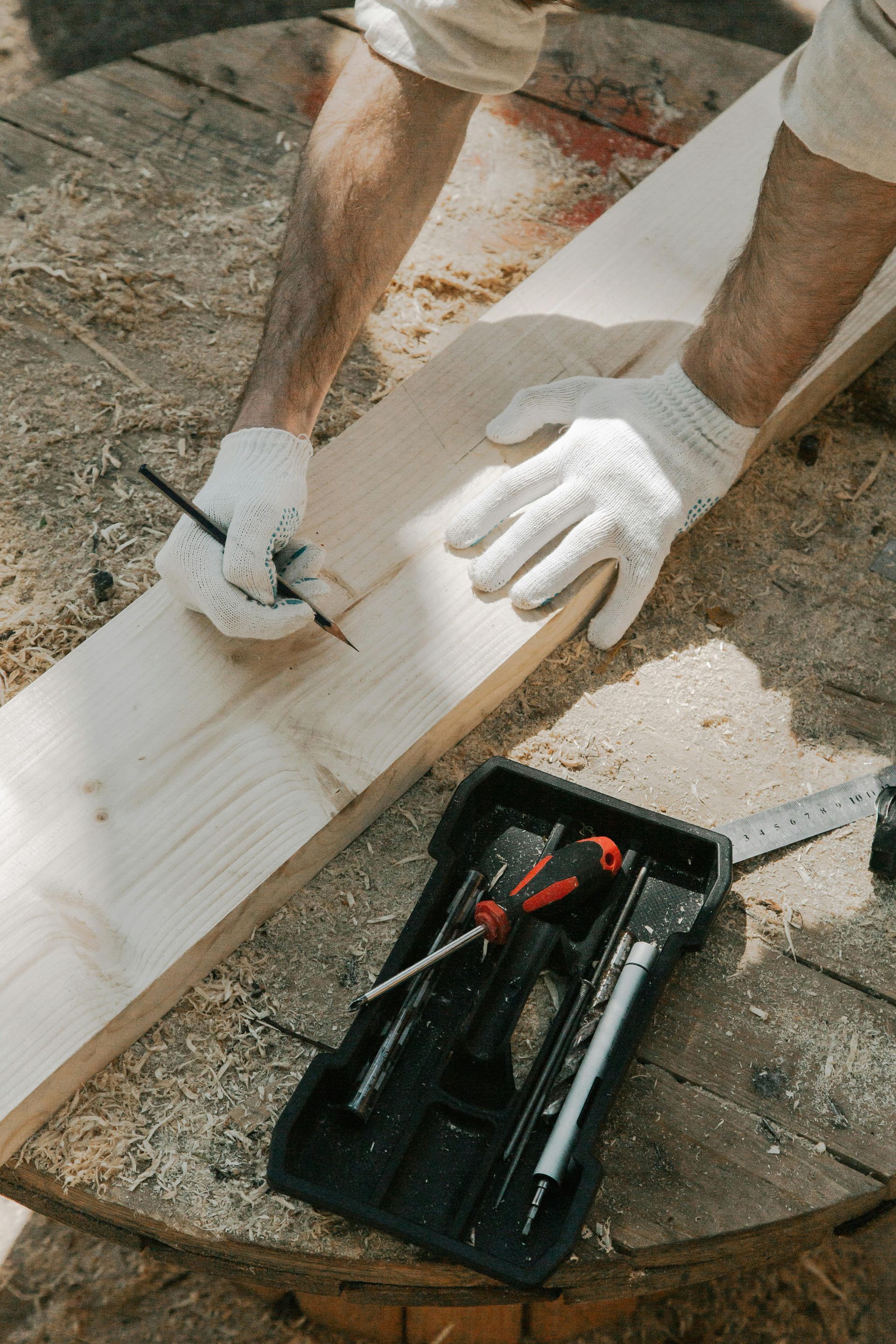 Person wearing gloves marks a measurement on a wooden plank with a pencil, tool set nearby.
