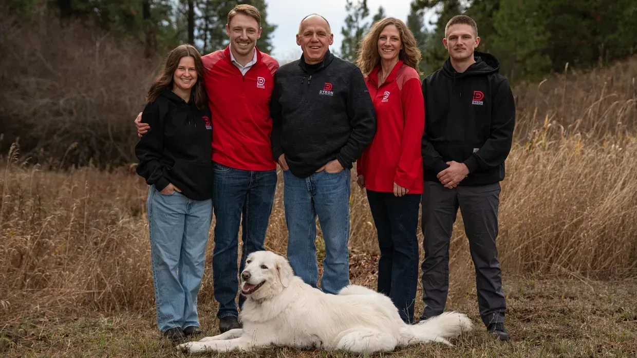 Family poses outdoors with a white dog. They wear red and black shirts, smiling, in a field.