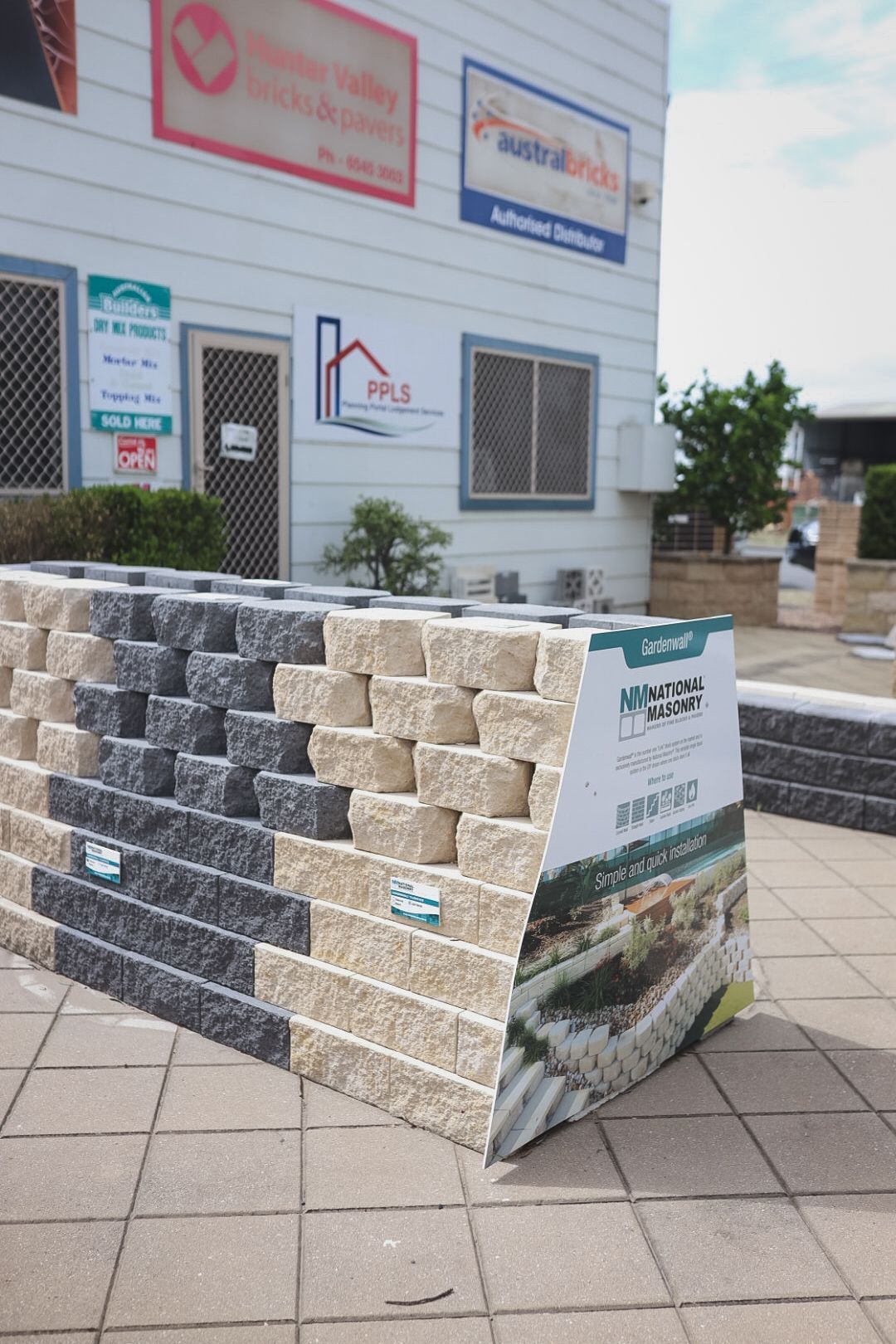 Stacked sample wall of gray and beige stone blocks displayed outdoors on a tiled sidewalk.— Hunter Valley Bricks & Pavers in Scone, NSW