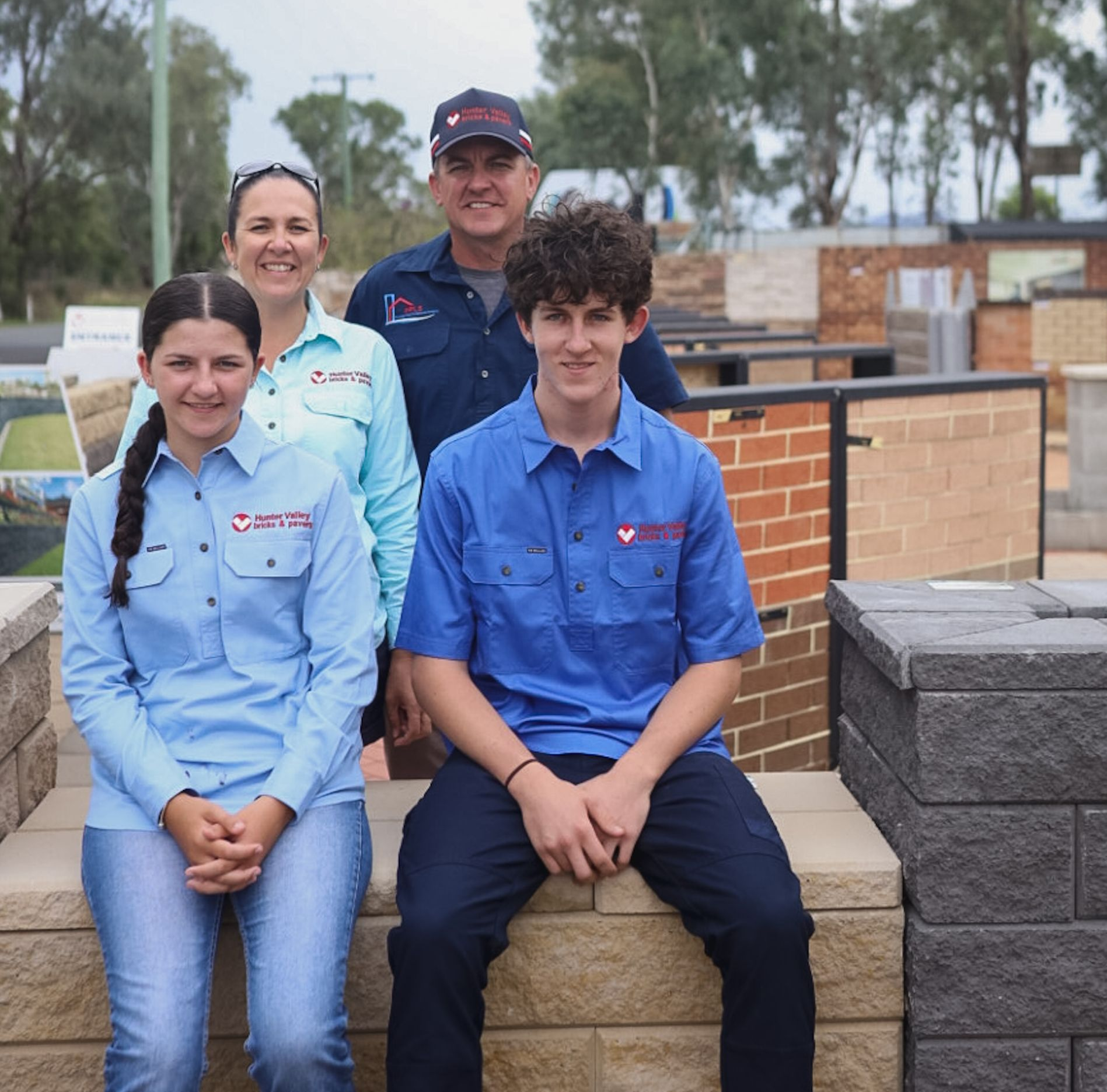 Four people stand and sit together at a masonry supply yard, wearing matching blue branded shirts.
