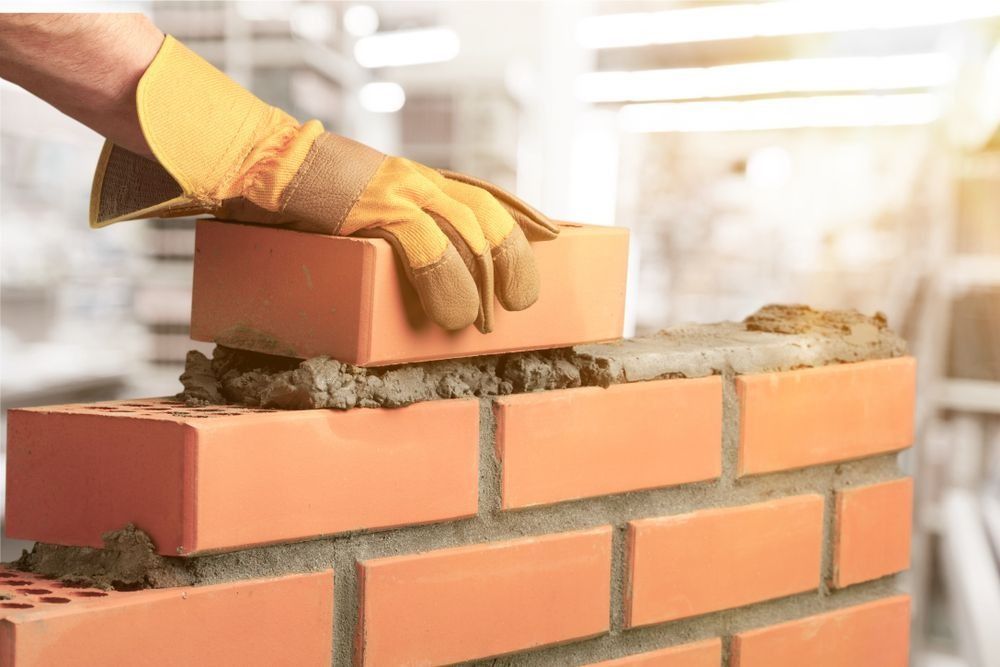 Hand in Work Glove Placing a Red Brick Onto a Mortar-covered Brick Wall — Hunter Valley Bricks & Pavers in Scone, NSW