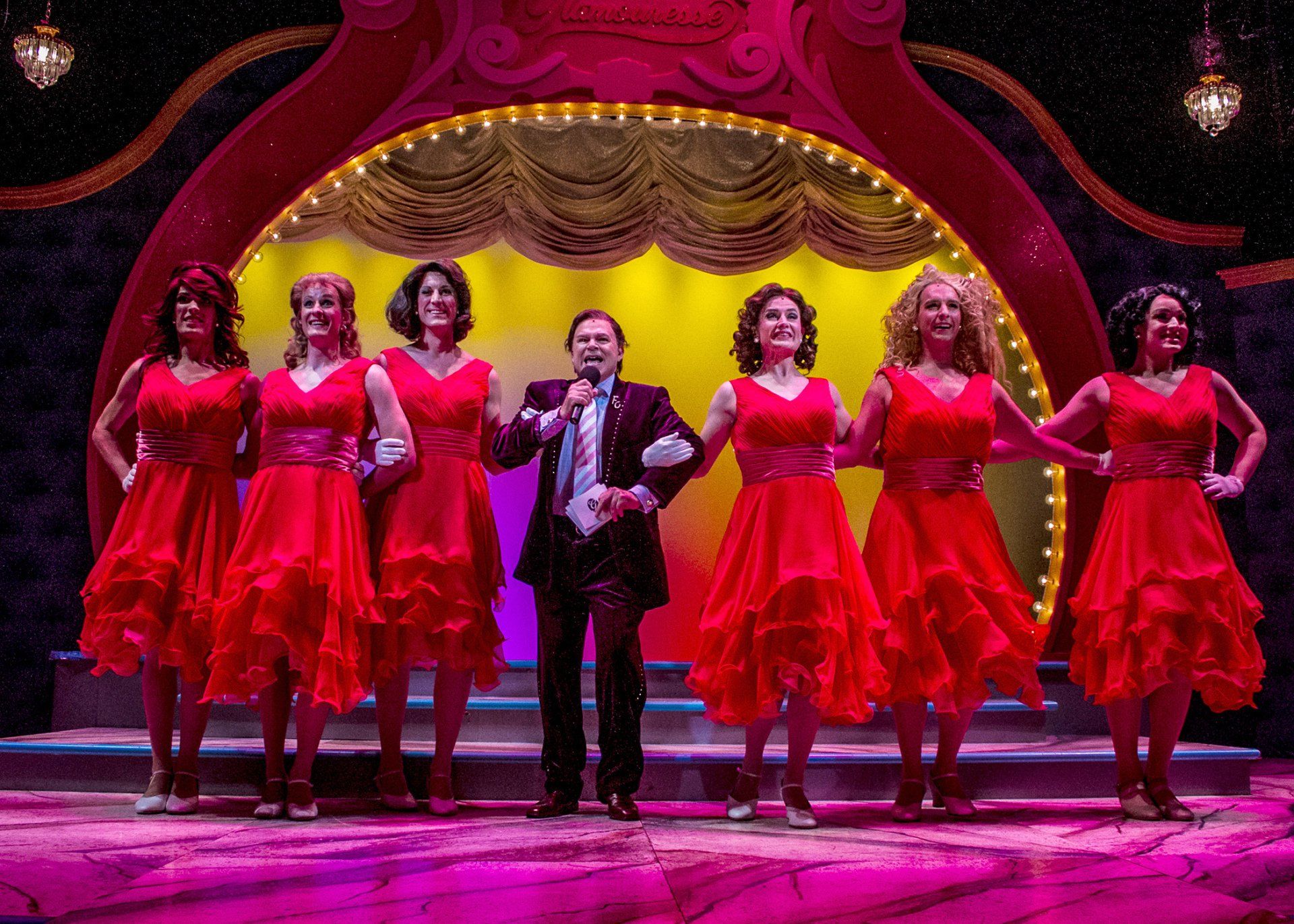 6 actors onstage wearing red dresses and one actor holding mic and wearing dark suit