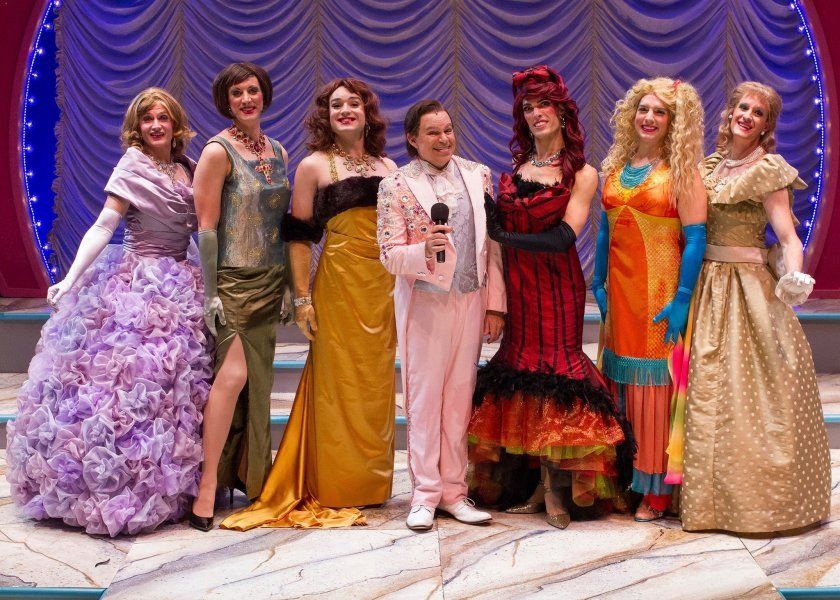 6 actors onstage wearing colorful dresses and one actor holding mic and wearing pink tux