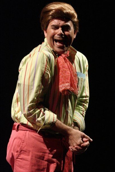 1 actor onstage making a silly face and wearing blonde wig, striped shirt, and pink scarf tie