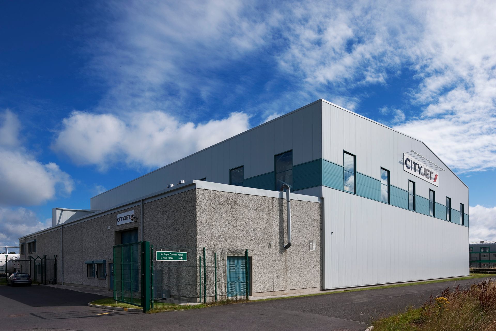 A large white building with a green stripe on the side is surrounded by a fence.