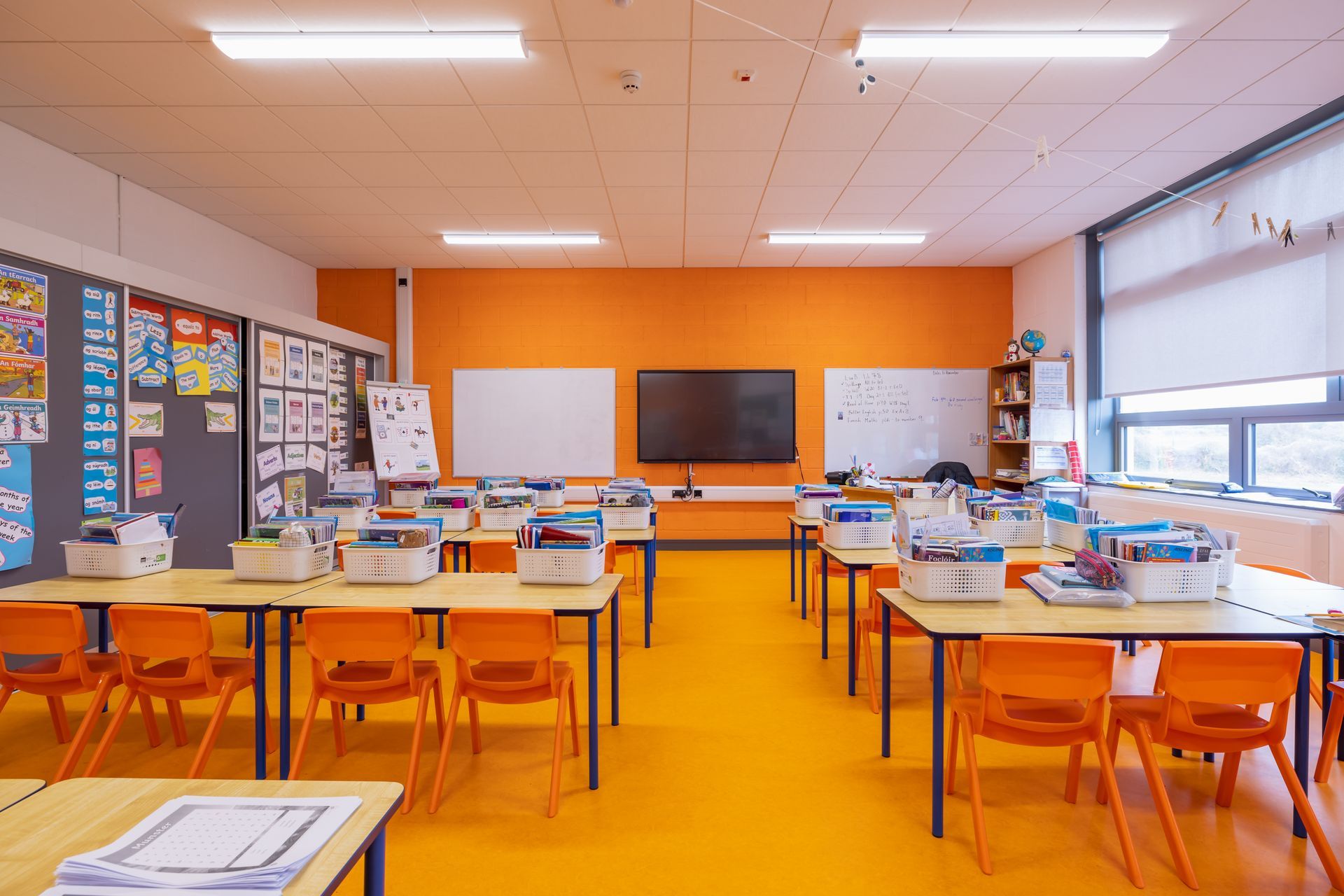 An empty classroom with orange tables and chairs and a large flat screen tv.
