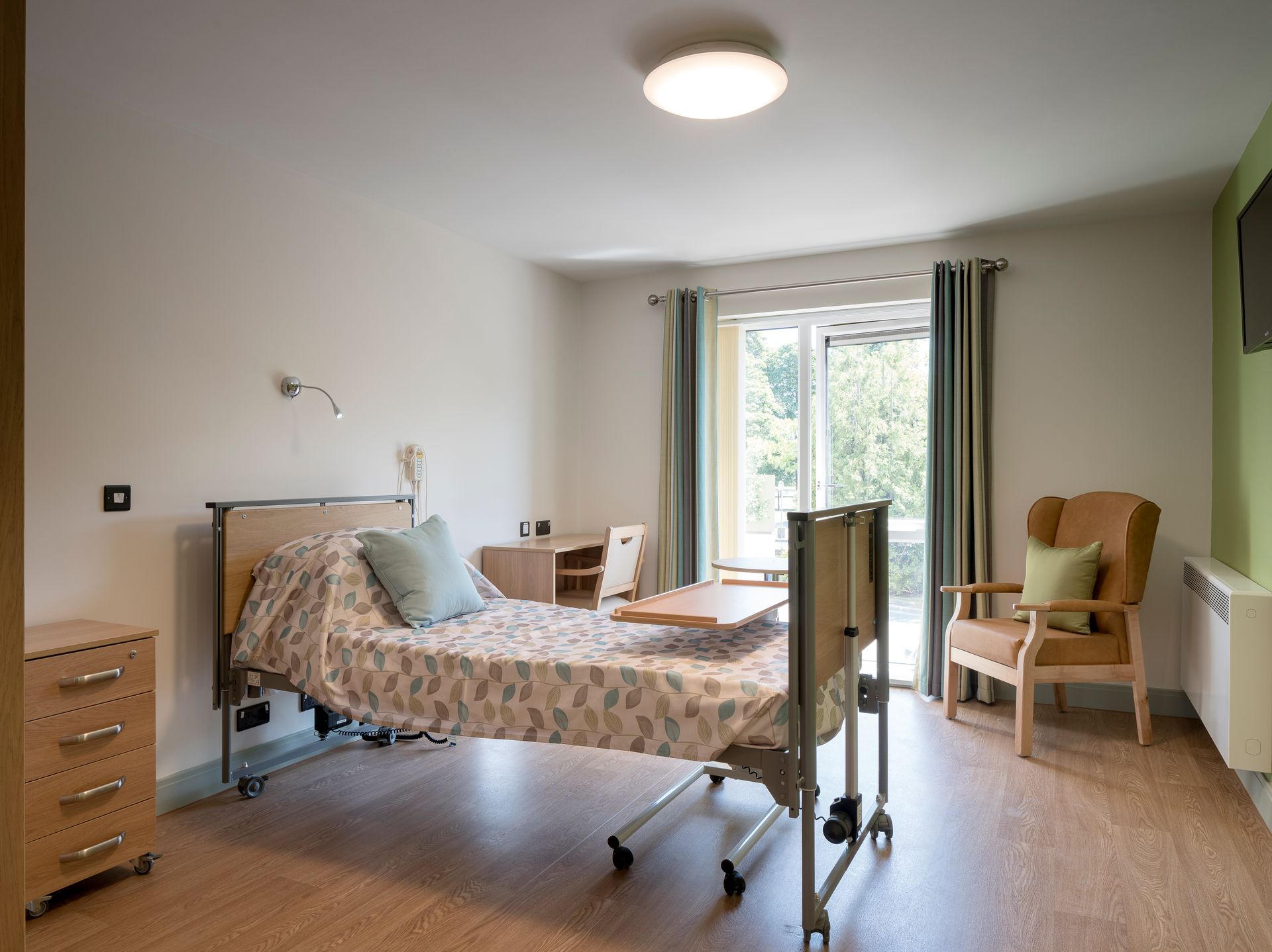 A hospital room with a bed , chair , desk and television.