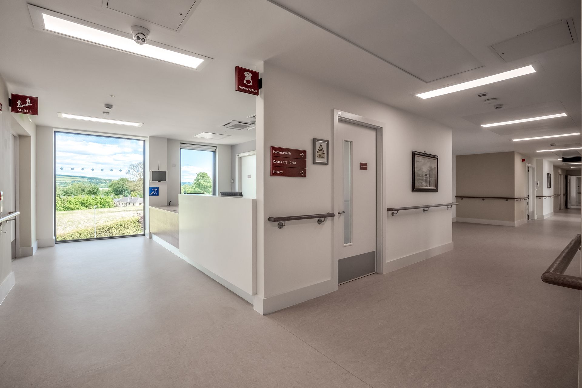 A long hallway in a hospital with a reception desk and a large window.