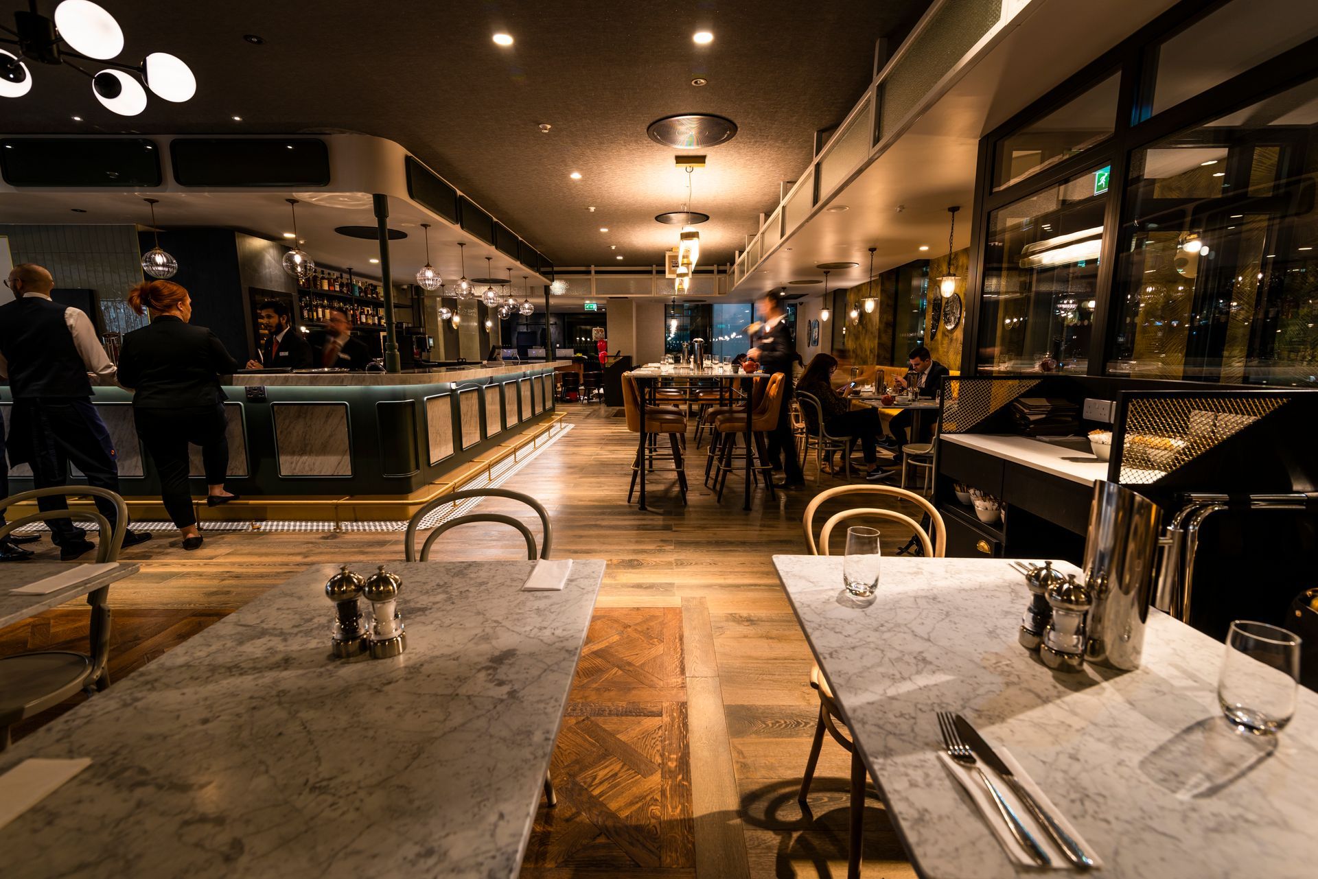 A restaurant with tables and chairs and people sitting at the bar.