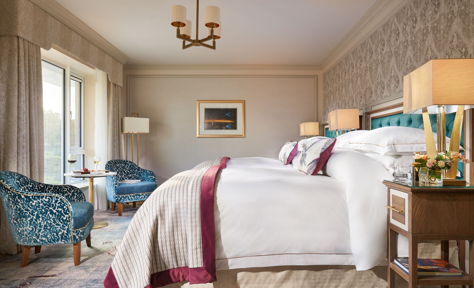 A hotel room with two beds , chairs , lamps and a chandelier.