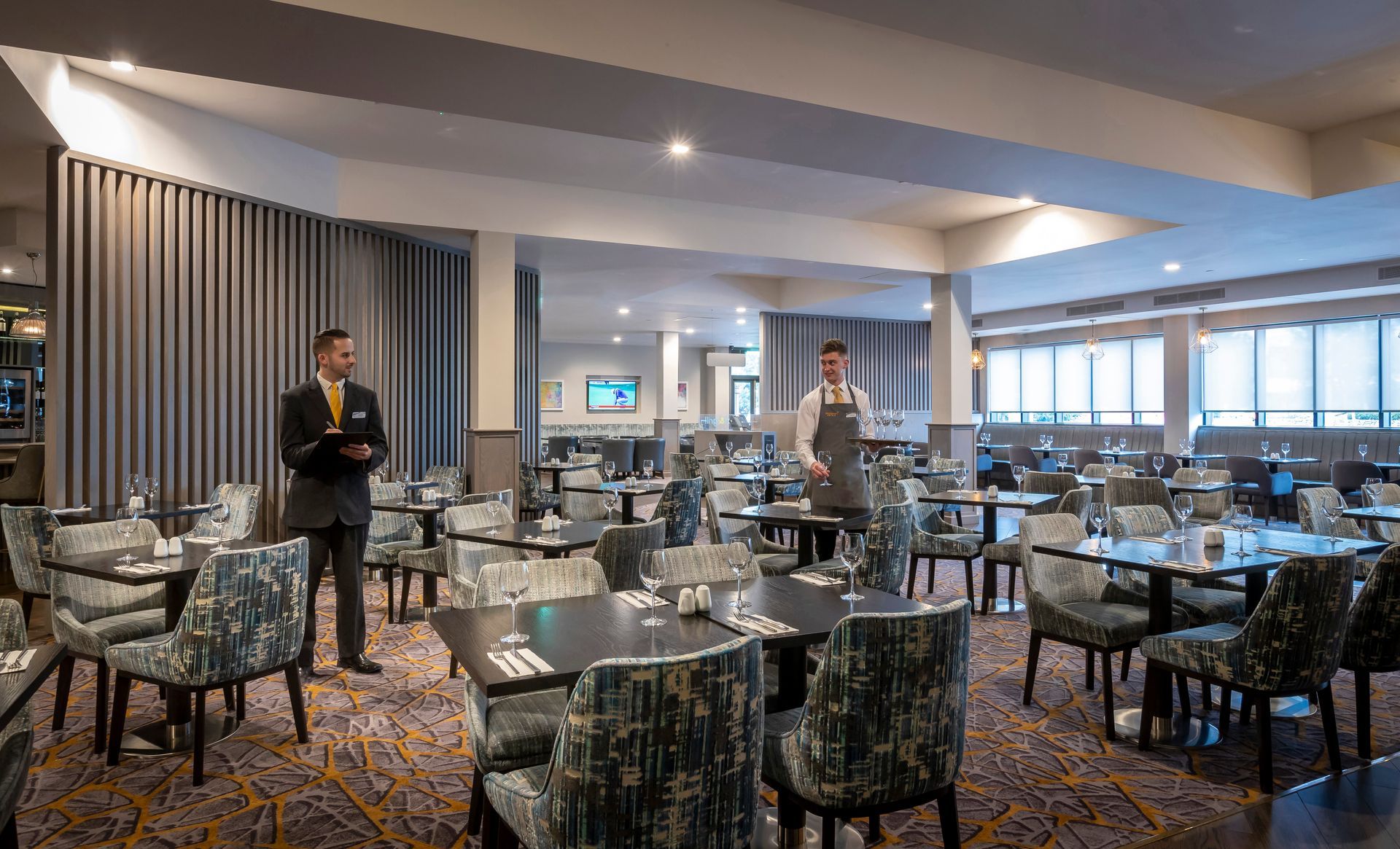 A restaurant with tables and chairs and two waiters standing in the middle of the room.