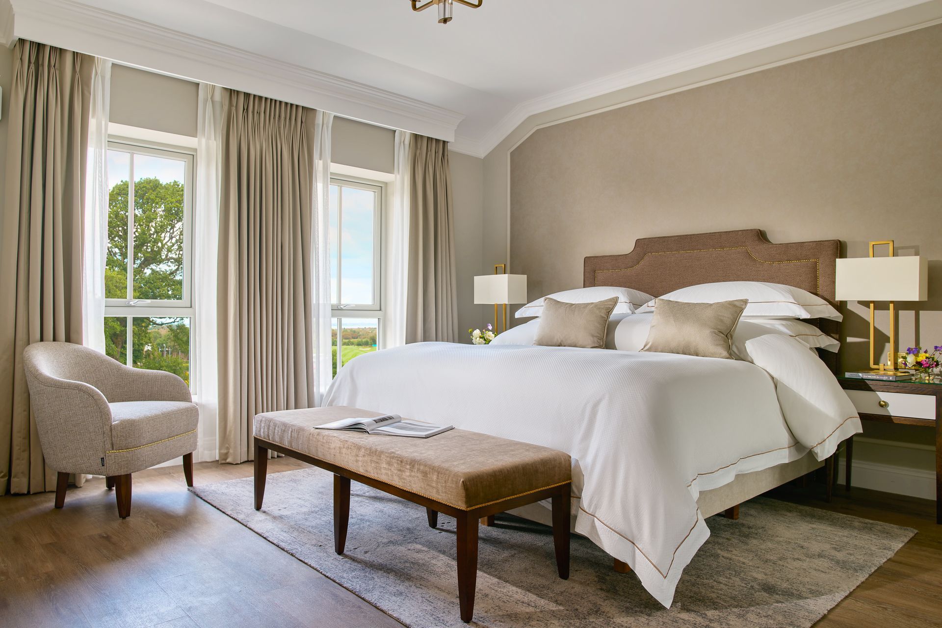 A hotel room with a king size bed , chair , bench and window.