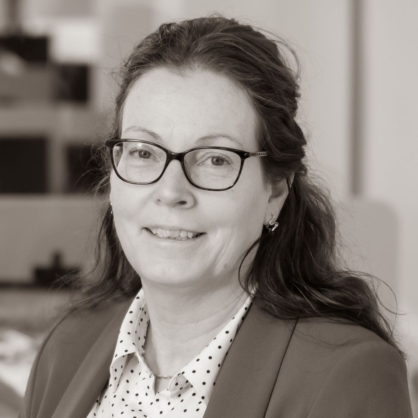 A woman wearing glasses and a polka dot shirt is smiling in a black and white photo.