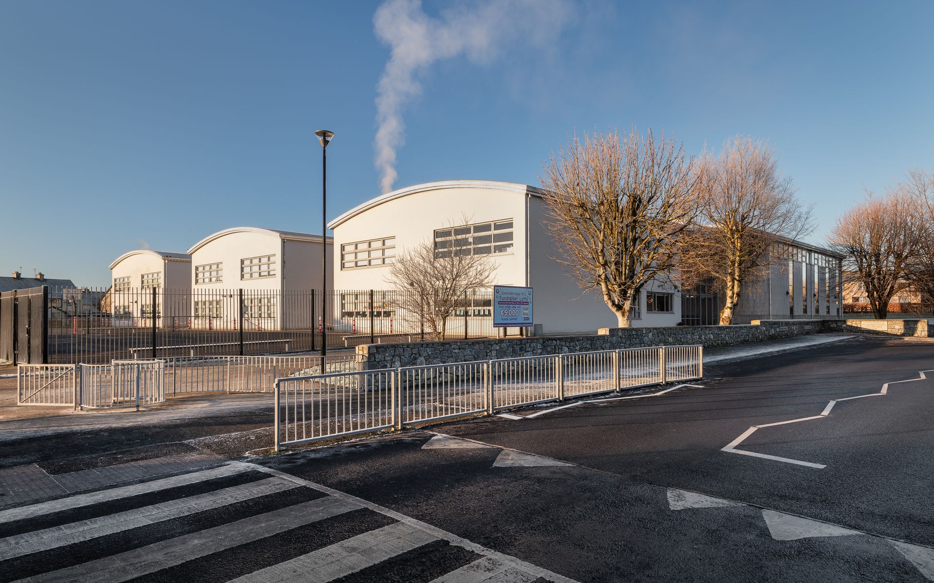 A large white building with smoke coming out of it is surrounded by trees and a fence.