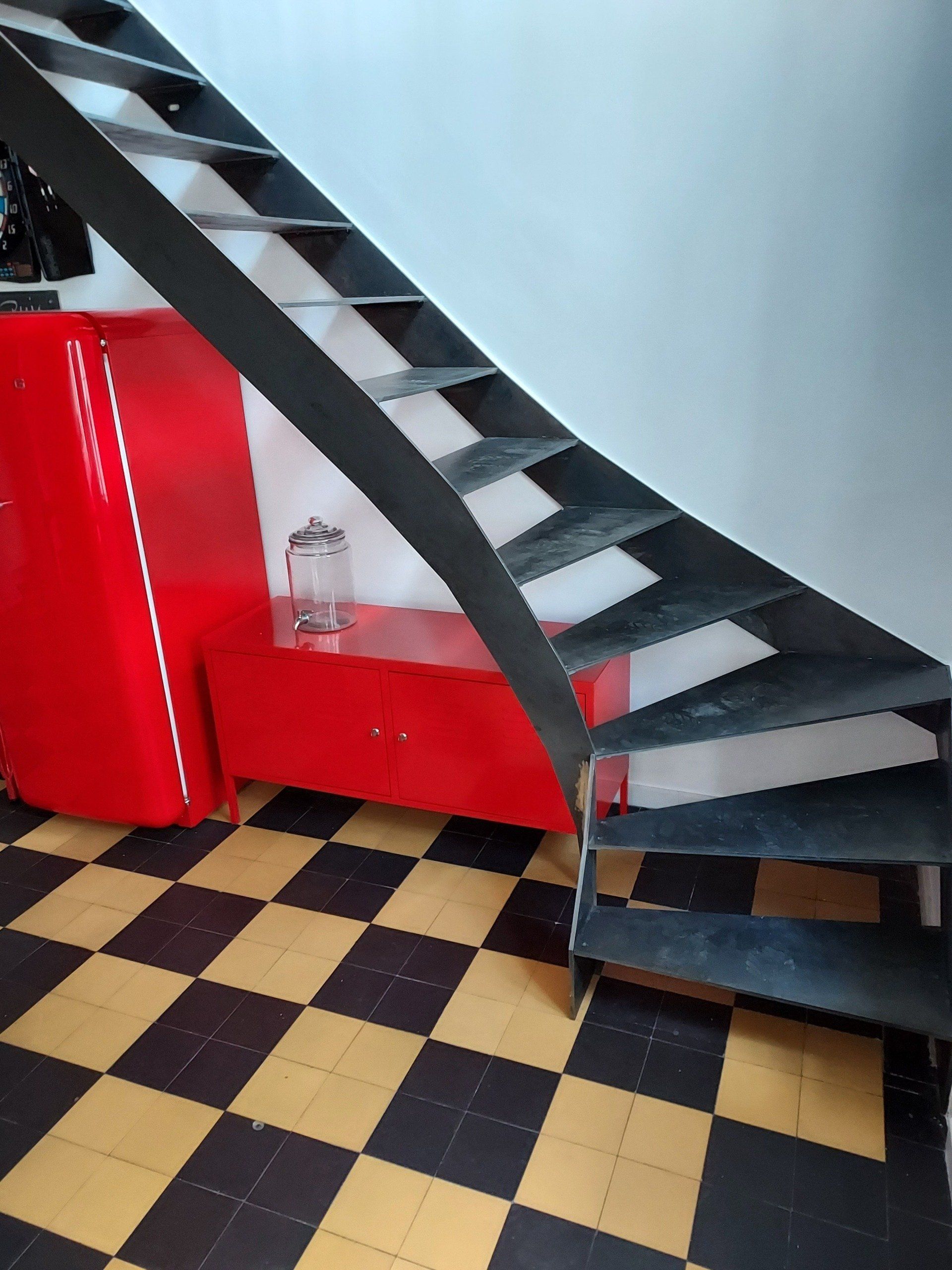 Un escalier en métal noir descend à côté d'un objet rouge sur un sol à carreaux jaune et noir.