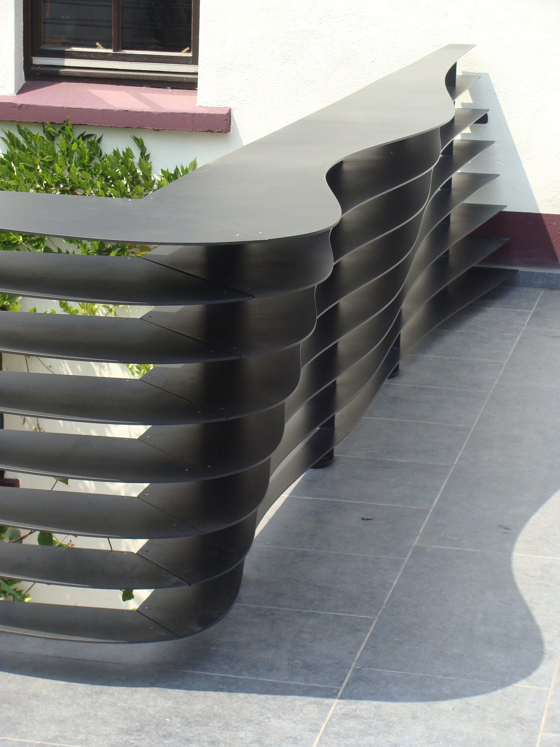Garde-corps extérieur en métal noir au design ondulé, contre un mur blanc et du carrelage gris.