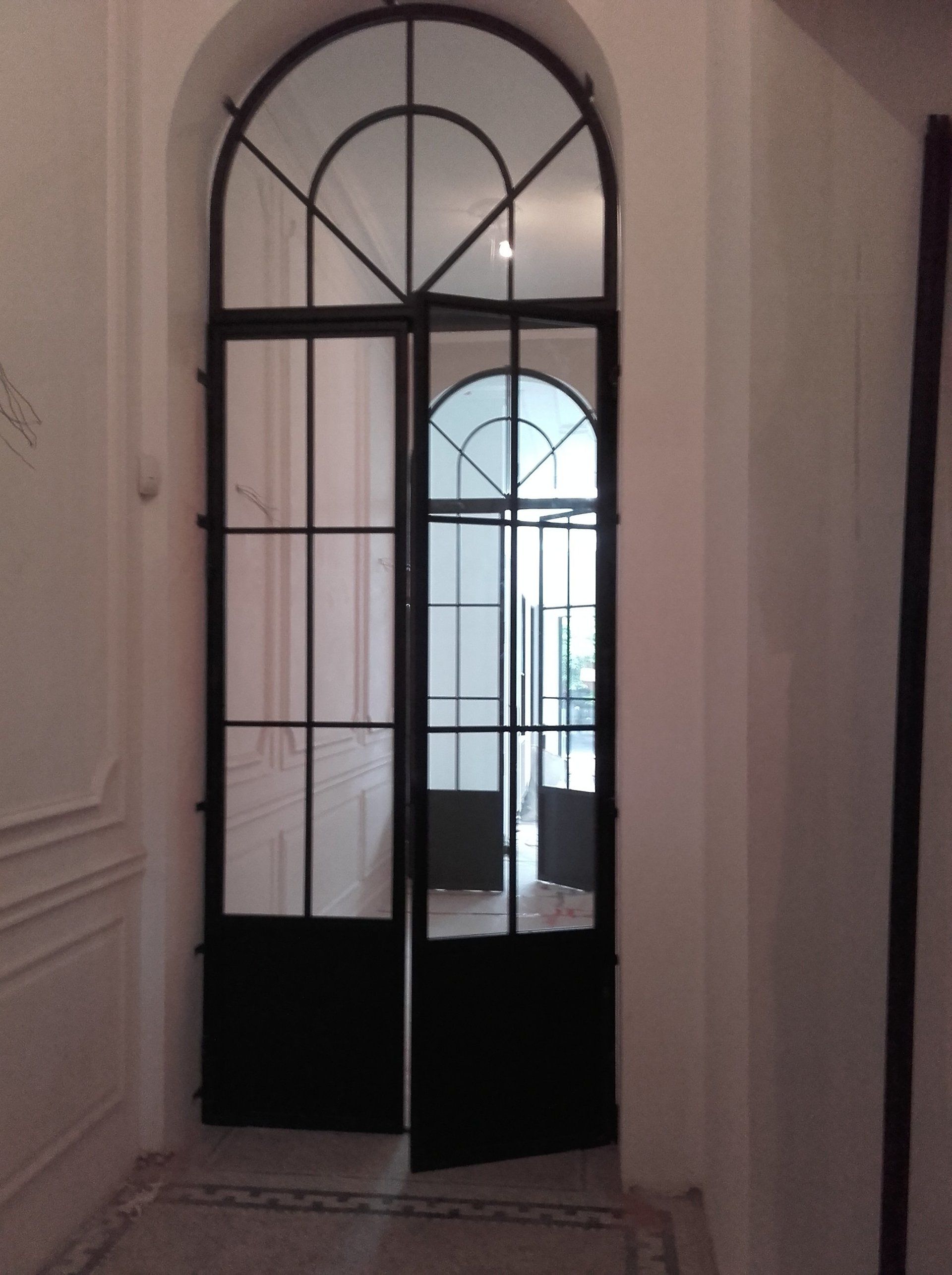 Portes en verre cintrées à cadre noir dans un couloir blanc avec un aperçu d'un espace intérieur.