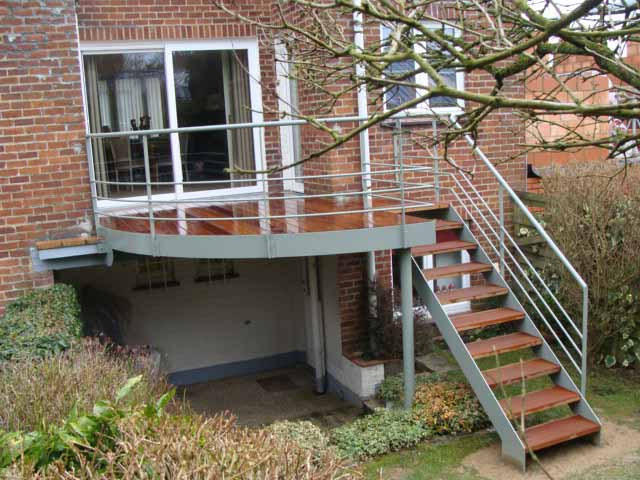 Bâtiment en briques avec balcon et escalier en métal. Terrasse, escalier et garde-corps en bois marron.