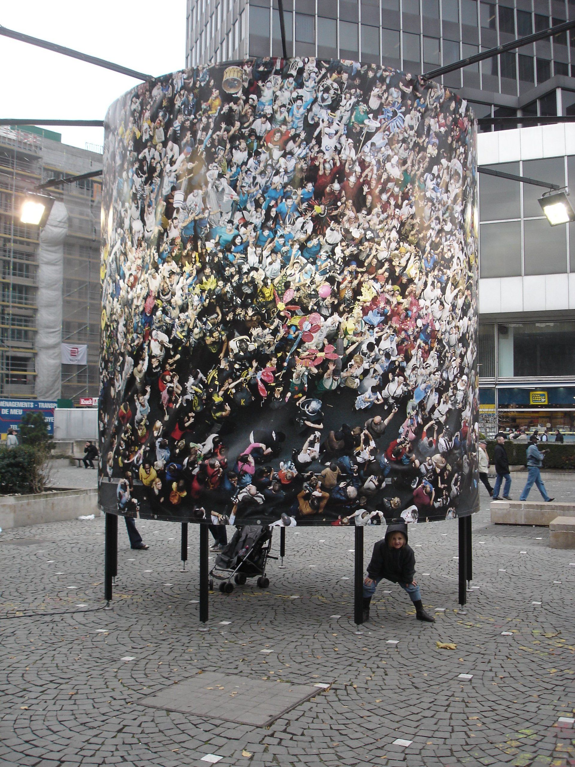 Grande installation artistique cylindrique sur pieds, sur une place publique, recouverte de fragments colorés