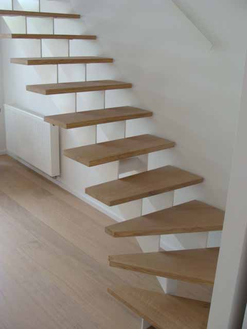 Escalier flottant en bois contre un mur blanc avec un radiateur, menant vers le haut depuis un plancher en bois clair.