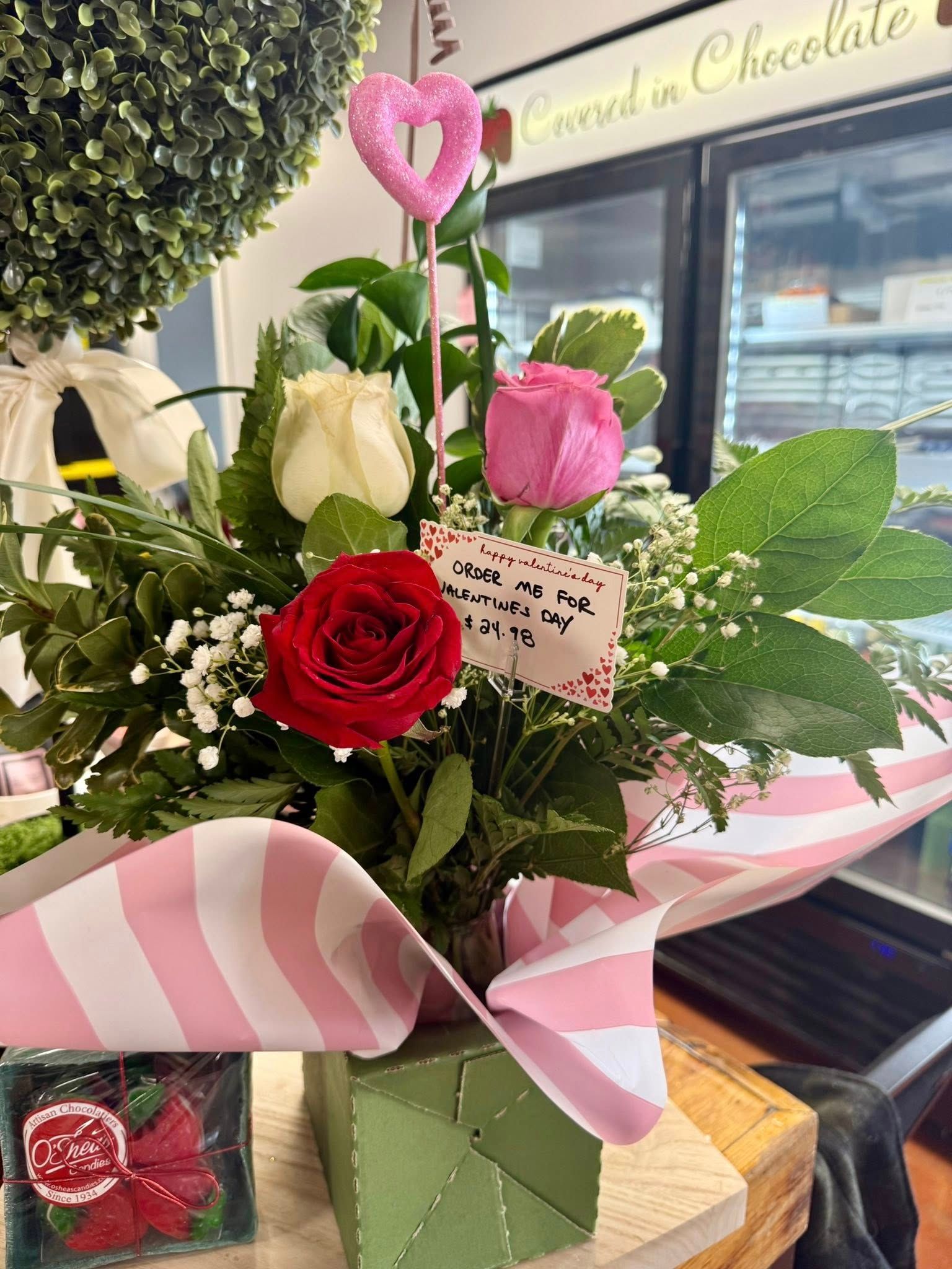 A floral arrangement with a red, white, and pink rose, greenery, and a heart pick in a green box with pink striped wrap.