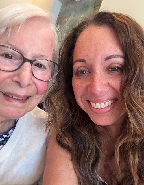 Two women smiling close to camera. Older woman wears glasses and cardigan. Younger woman has wavy hair.