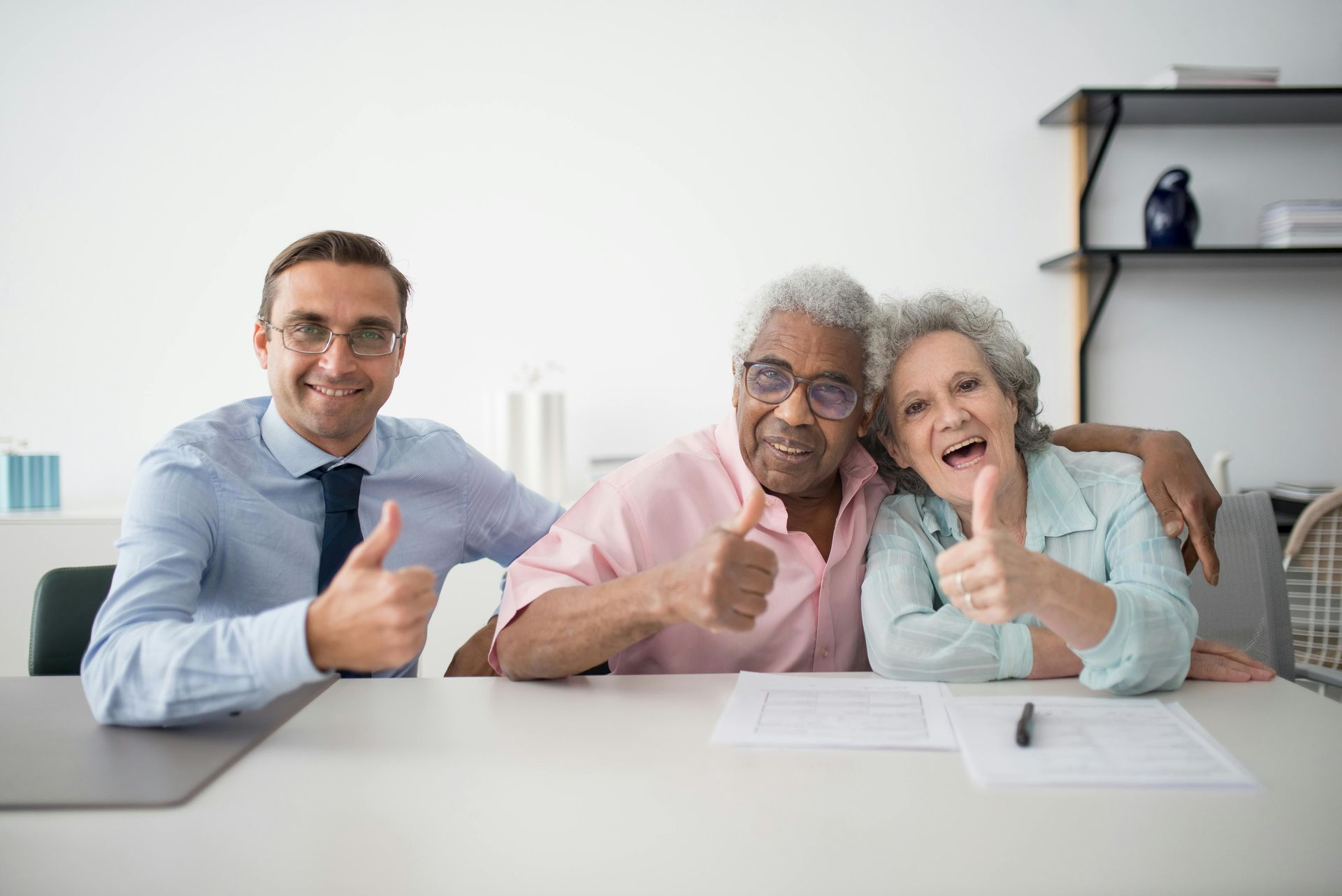 Man and older couple giving thumbs up at a table. Papers and pen are visible.