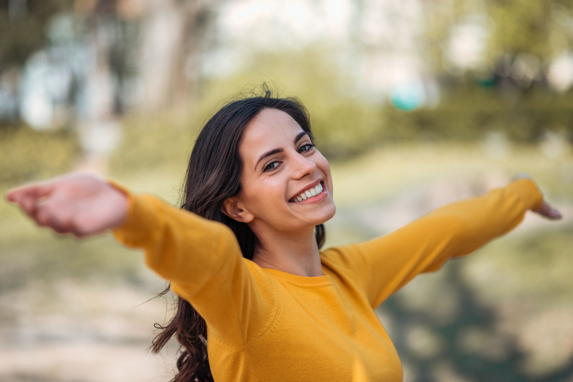 A woman in a yellow sweater is smiling with her arms outstretched.