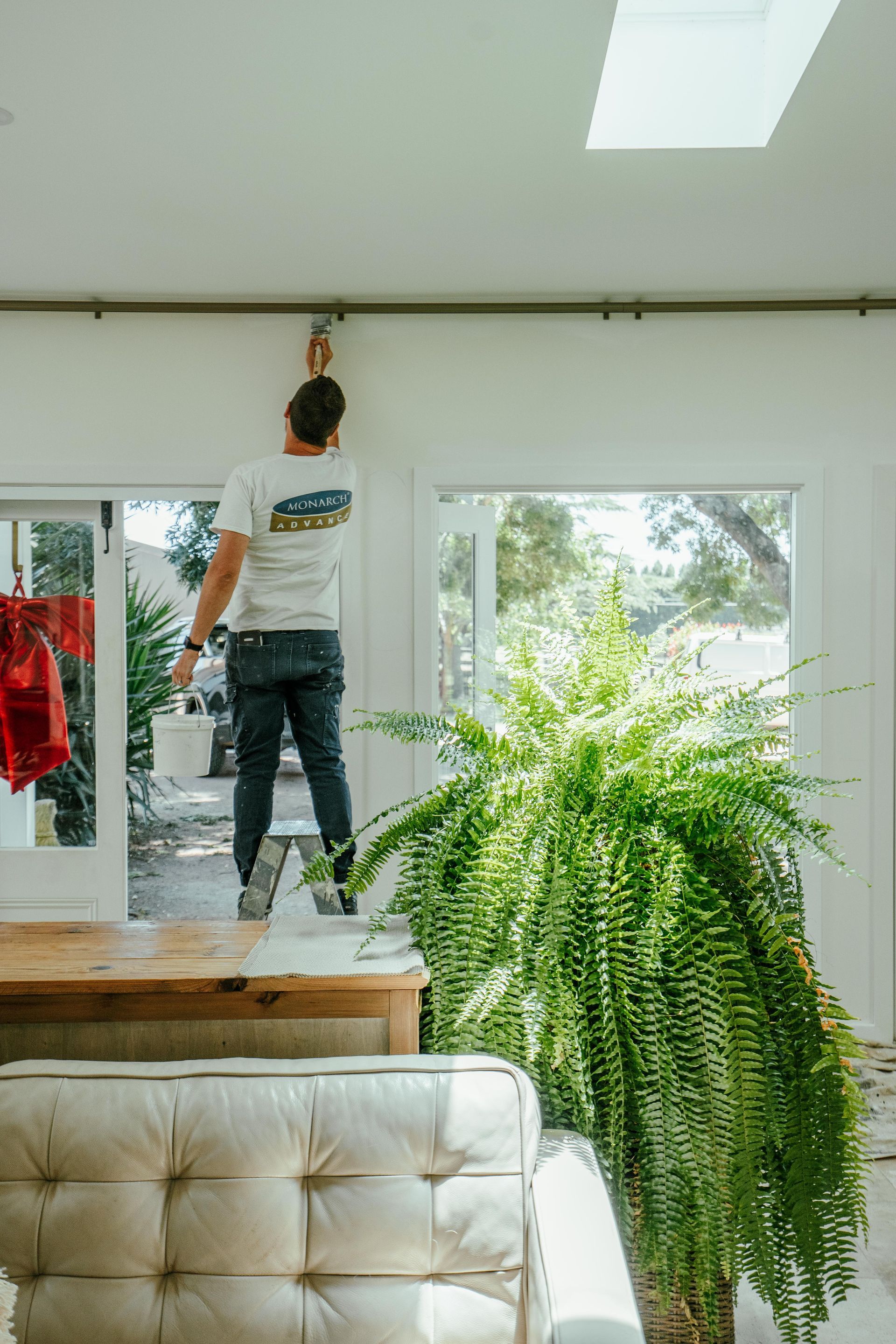 Man on a table paints a ceiling fixture. Bright, indoor space with large fern and open windows. — Mittagong Painting in Welby, NSW