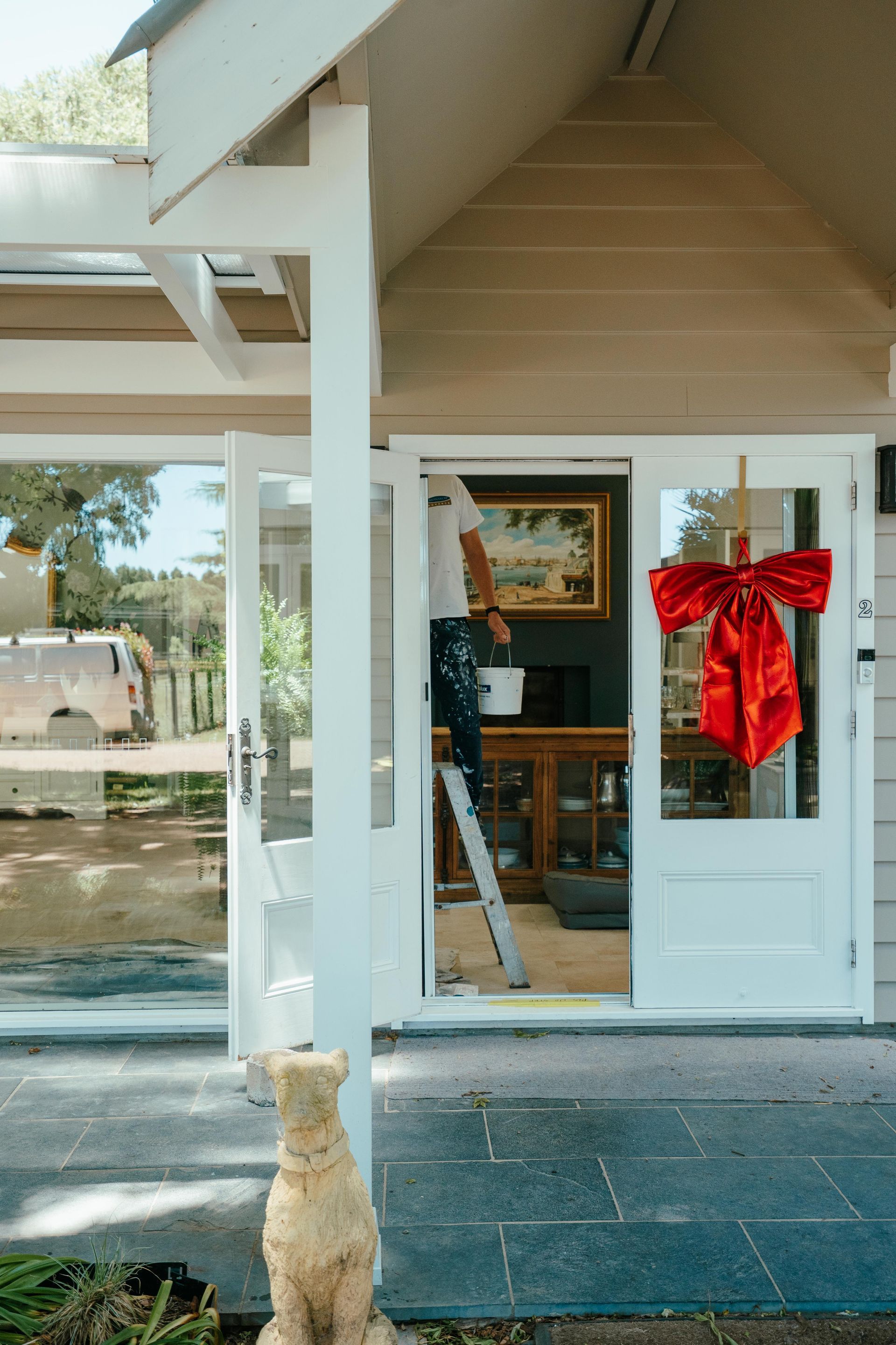 Person on a ladder painting a white doorway. A red bow decorates the door. Building exterior. — Mittagong Painting in Welby, NSW