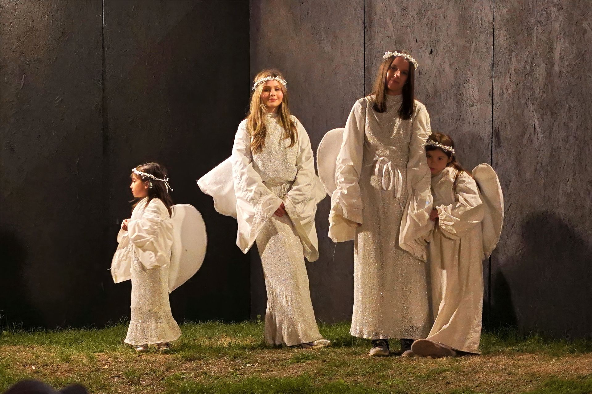 Four people in flowing white costumes stand against a dark wall at night.