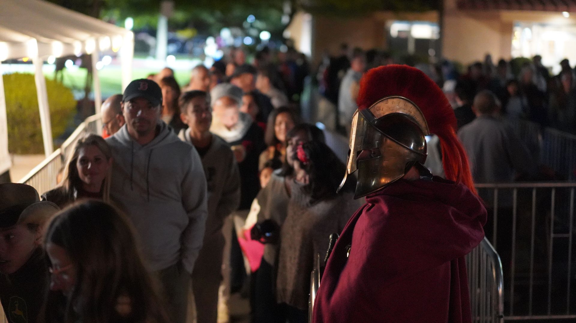 Crowd gathered outdoors at night near a lit walkway, with a person in a red helmet in the foreground
