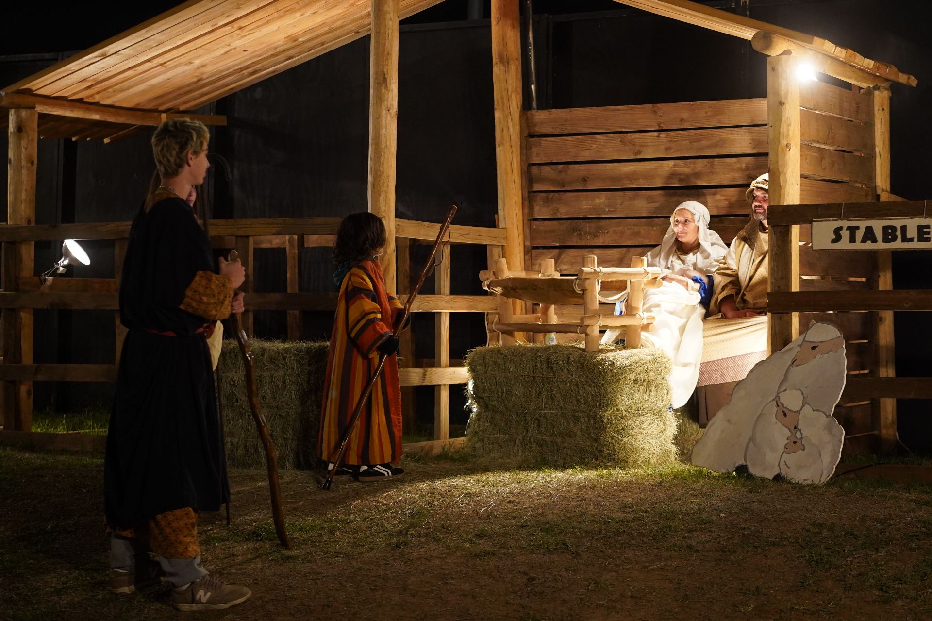 Nighttime stable scene with three people and a small animal near hay bales under warm lights.
