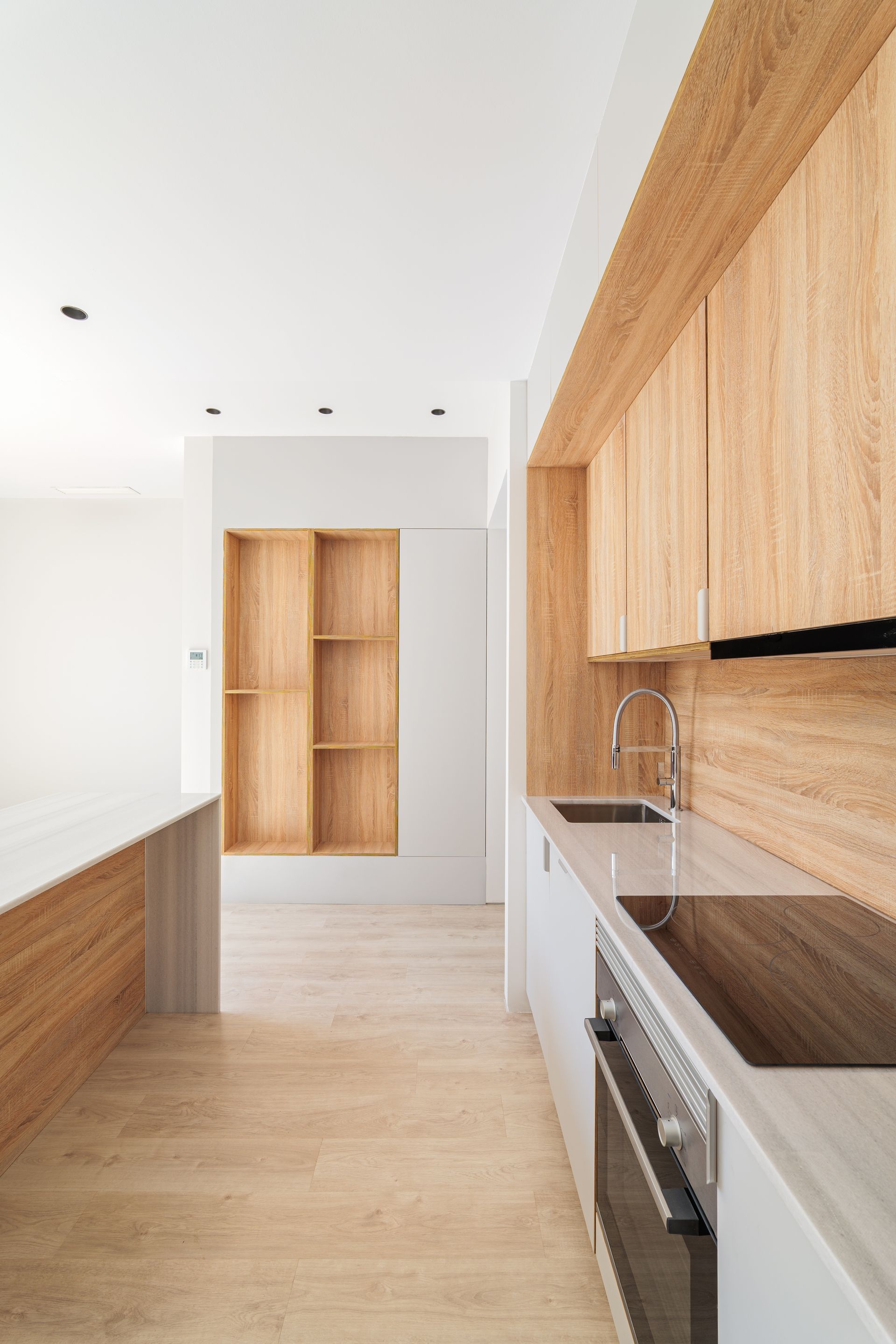 Narrow modern kitchen with light wood cabinets, white walls, and a built-in oven along the right side.