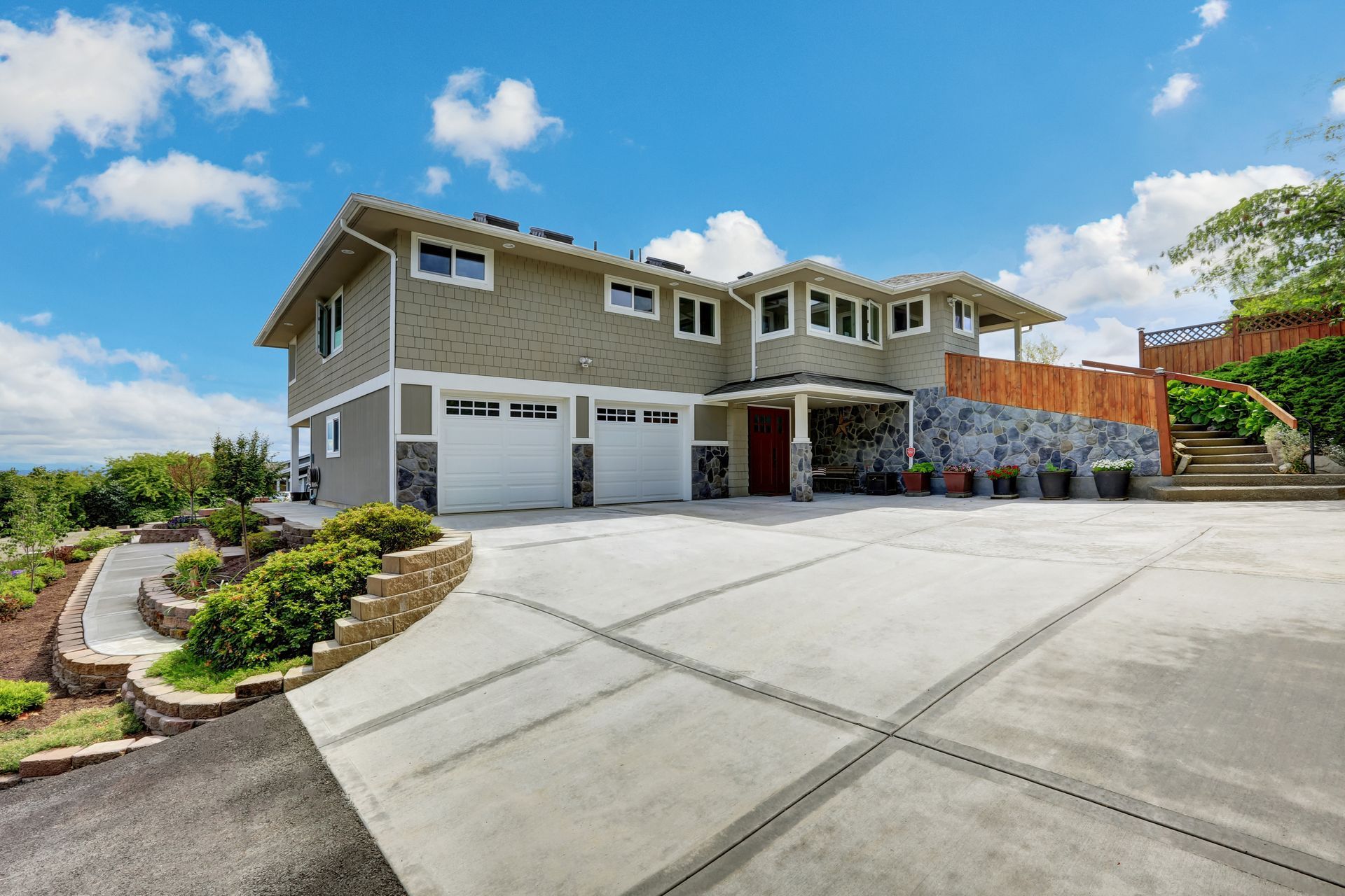 Large two-story suburban house with beige siding, garage doors, and a wide concrete driveway under a blue sky