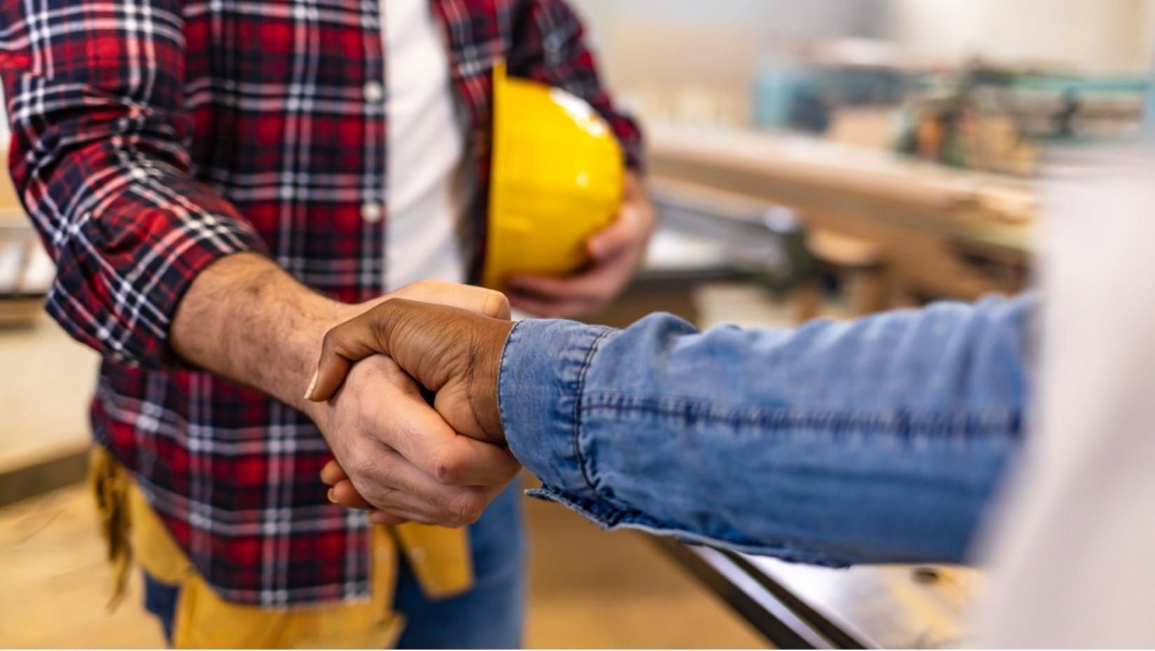 Two people shake hands, one holding a yellow hard hat in a construction setting.