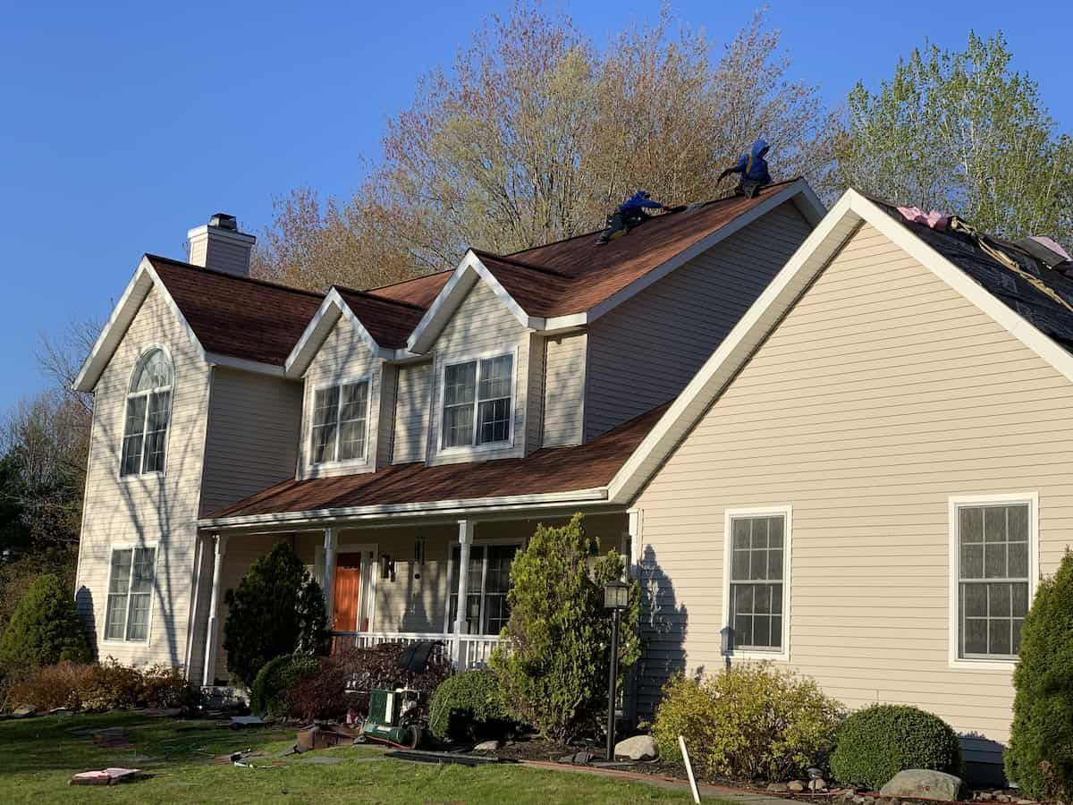 Two-story beige house with red roof, white trim, and a front porch in a sunny suburban yard