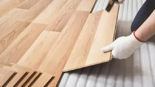 Gloved hands laying wood planks over a corrugated metal surface, installing flooring