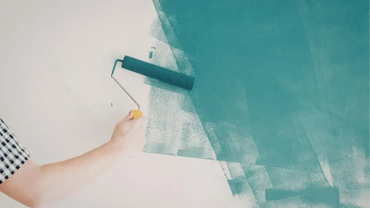 A hand using a paint roller to apply teal paint on a white wall.