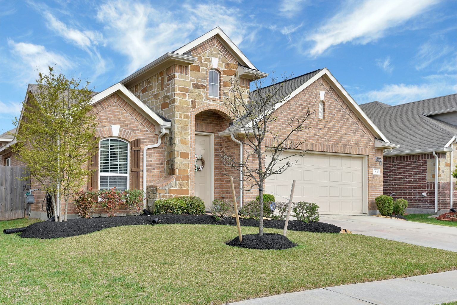 Brick suburban house with a two-car garage, manicured lawn, and young trees under a blue sky