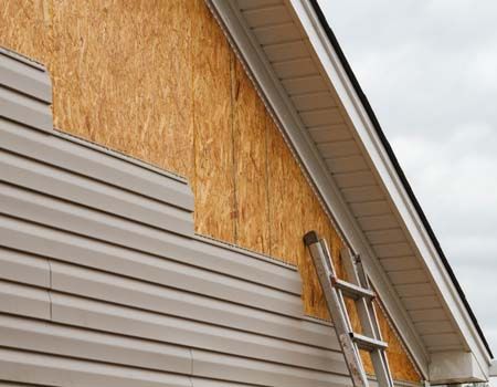 Exterior house wall with siding removed, exposed plywood sheathing, and a ladder leaning against the roofline