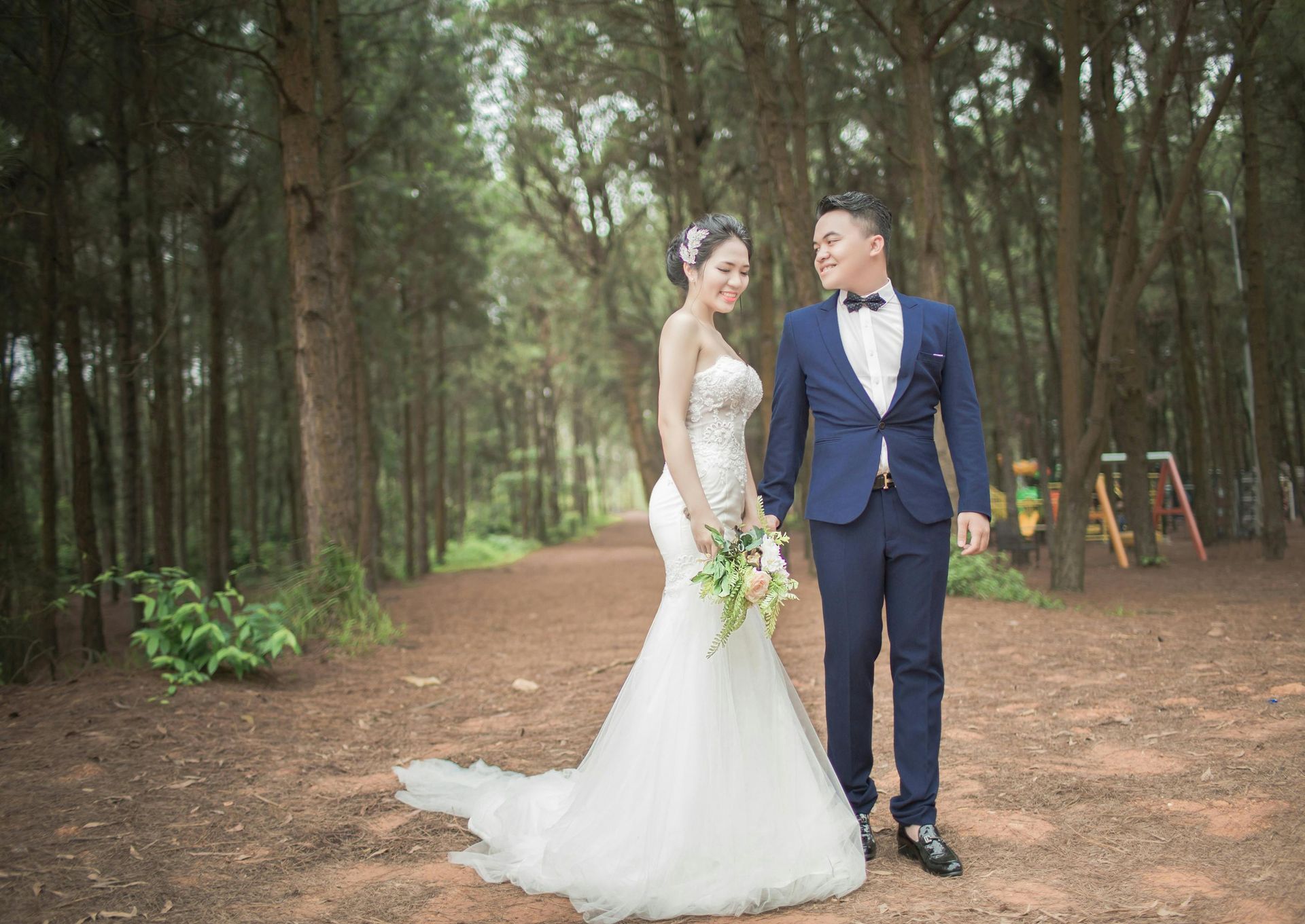 A bride and groom are walking through a forest holding hands.