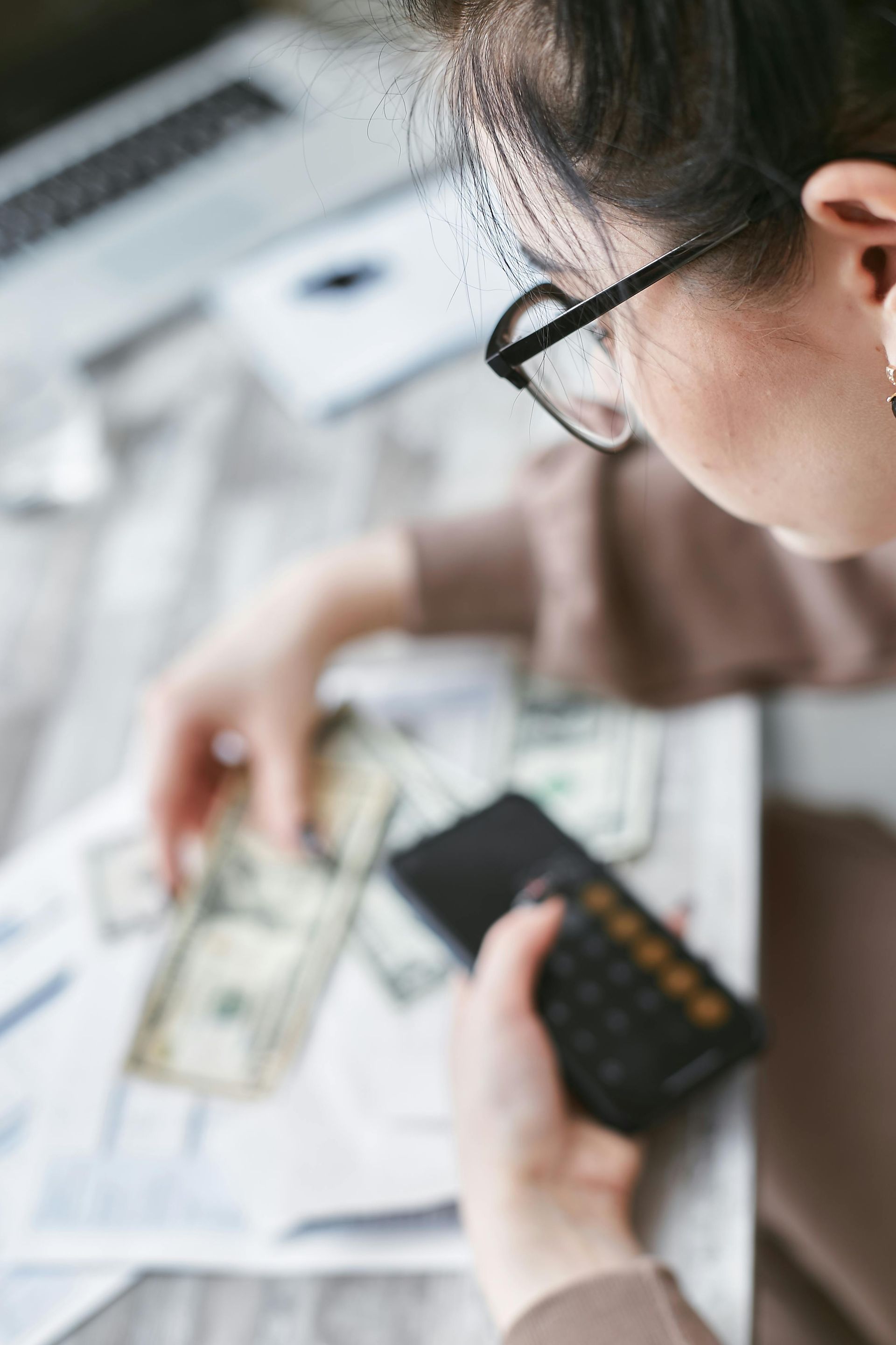 A person wearing glasses uses a calculator while sorting through US paper currency and documents on a desk.