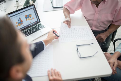 A person in a business suit points to a document while consulting with another person at an office desk with a laptop.