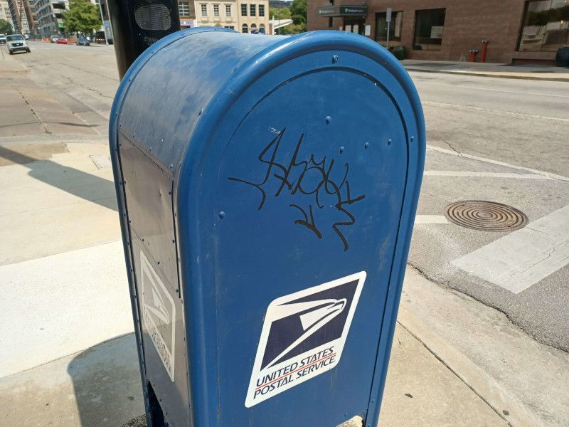 A blue mailbox on the side of the road has graffiti on it