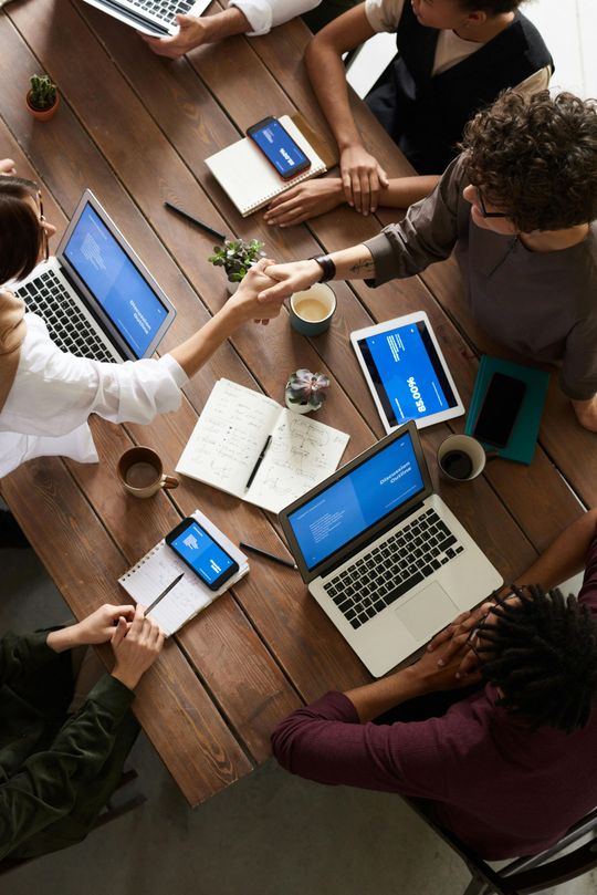 People at a wooden table in a meeting, shaking hands. Laptops, tablets, and phones are visible.
