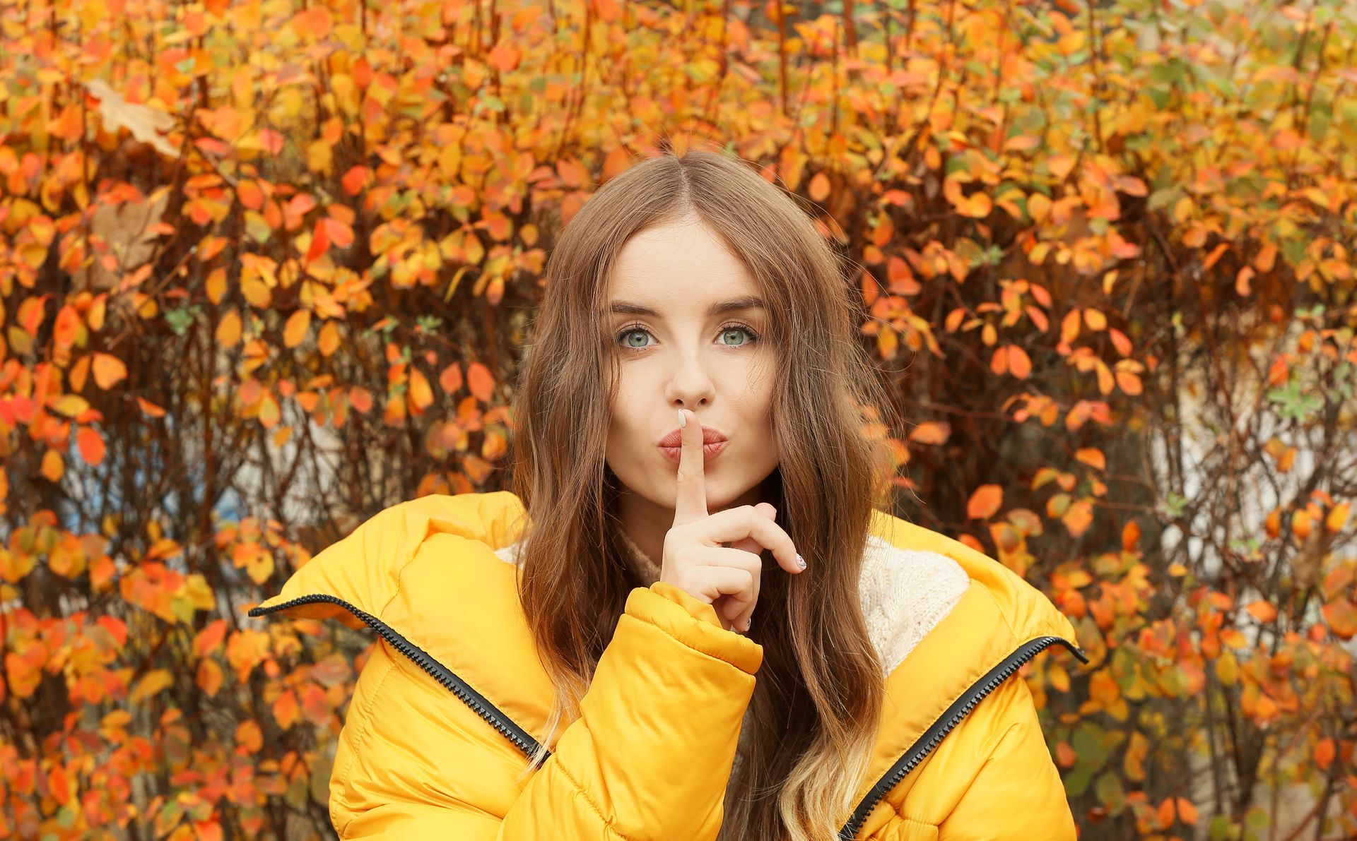 Woman with long brown hair in a yellow jacket holds finger to her lips in front of autumn leaves.