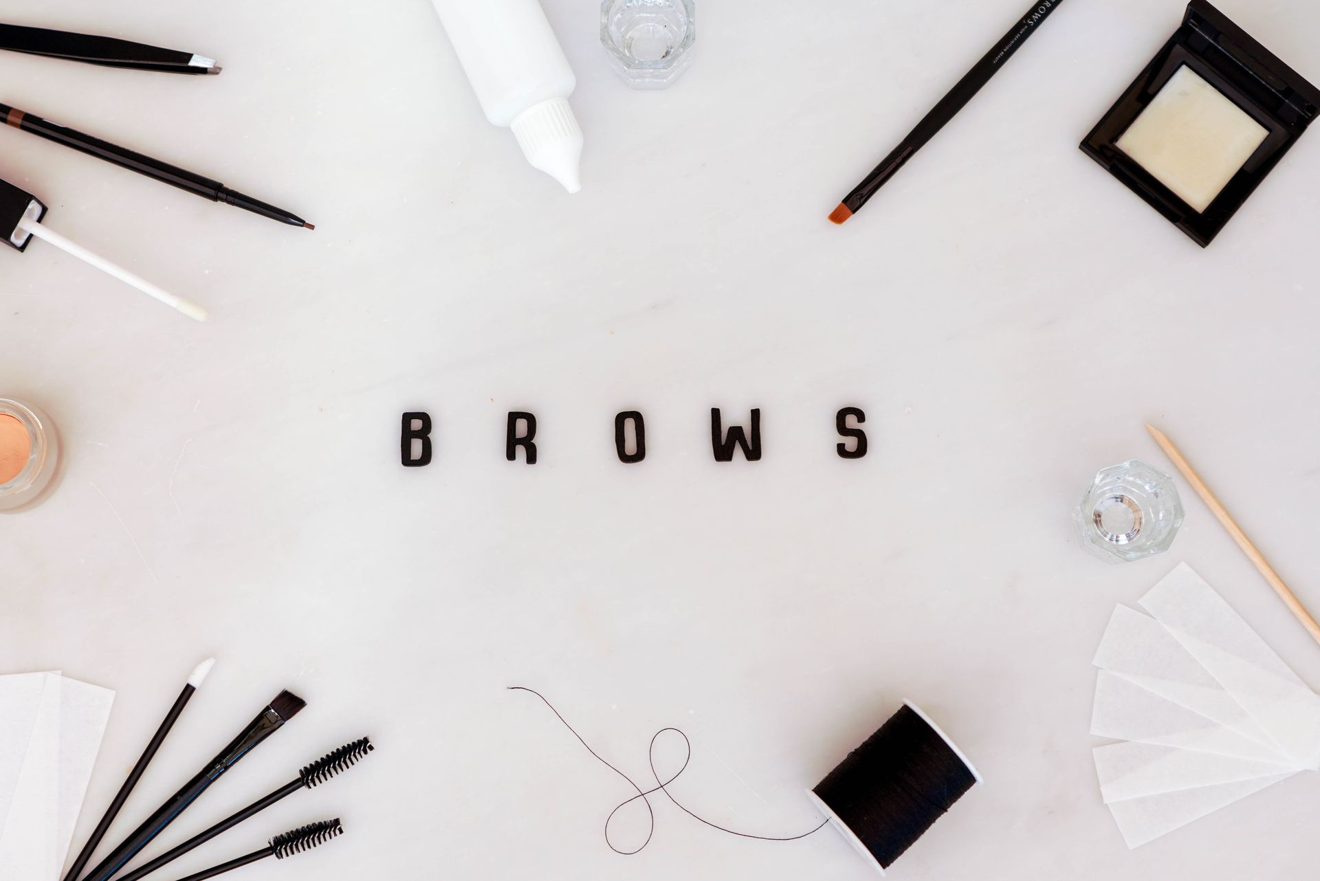 Cosmetics arranged around the word BROWS. Includes brushes, pencils, and other brow grooming supplies on a white surface.