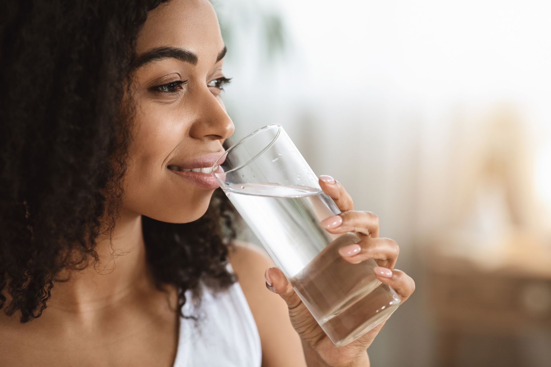 A woman is drinking a glass of water. She has clear and healthy looking skin.