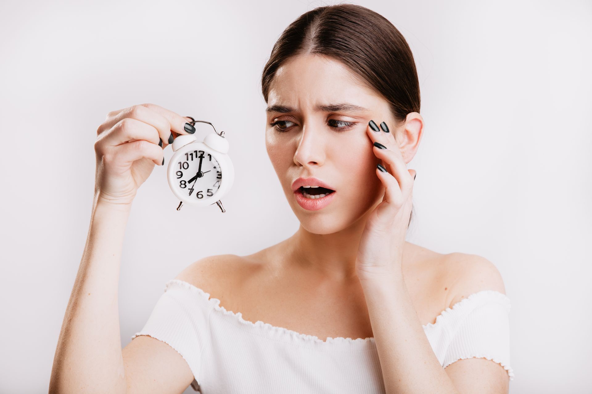 A woman is holding an alarm clock in her hand and rubbing her eyebrows.